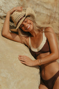Woman in a bikini and straw hat leaning against a stone wall.