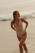 Woman in a beige swimsuit standing on a beach with waves in the background